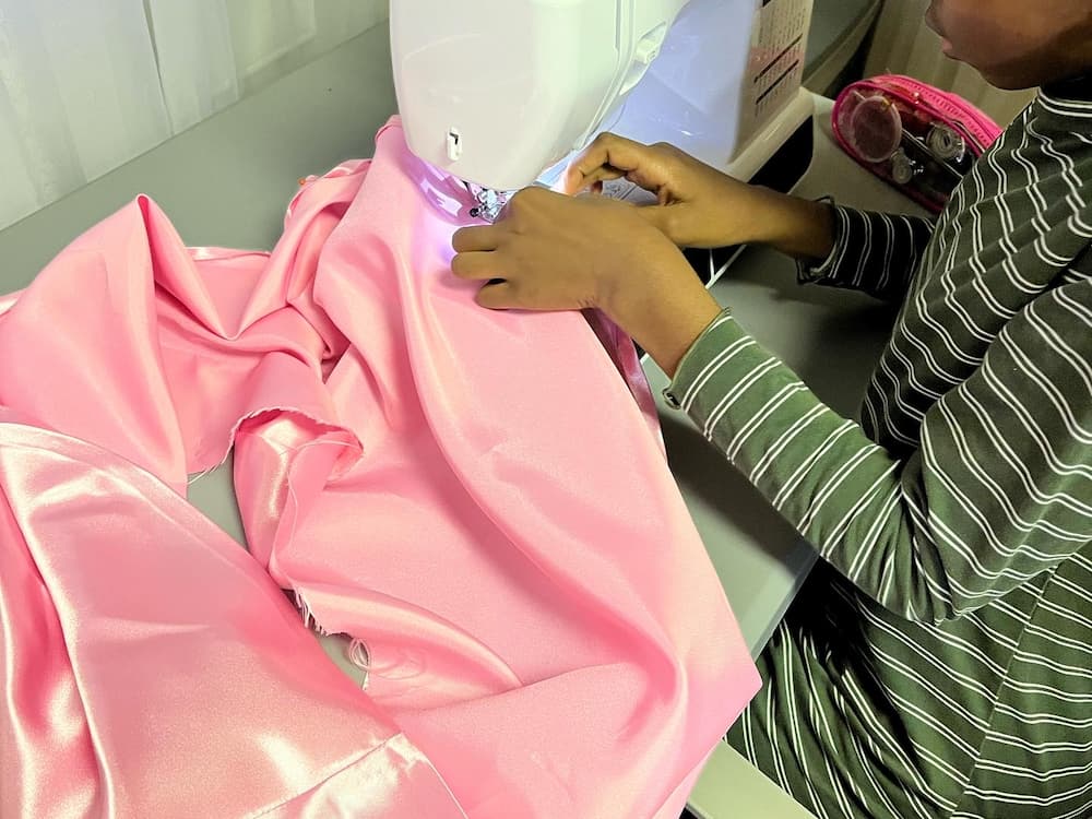 Close-up of a young student sewing a large piece of shiny pink fabric using a sewing machine.