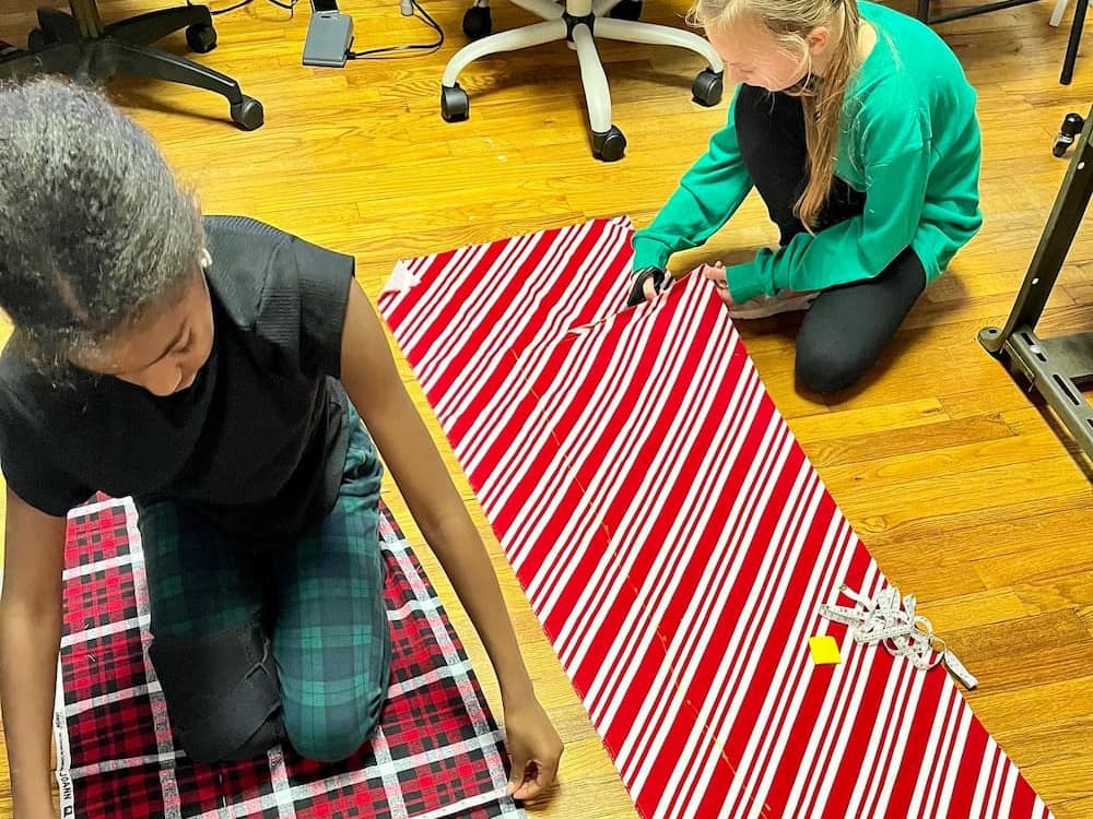 Two girls working on patterned fabric pieces, measuring and cutting material on a hardwood floor during a sewing project.