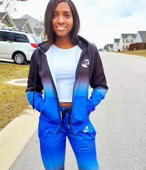 Brianna Michel standing outdoors in a blue and black tracksuit, smiling confidently in a suburban neighborhood.
