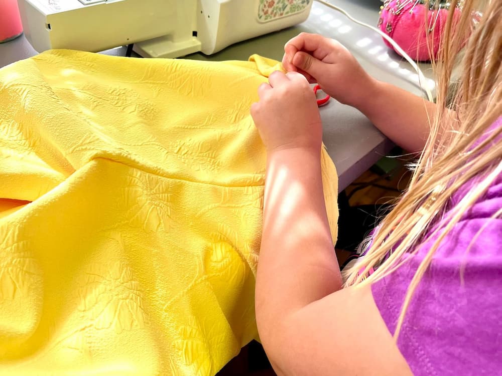 Child sewing a vibrant yellow fabric by hand at a table, with a sewing machine and pincushion nearby.