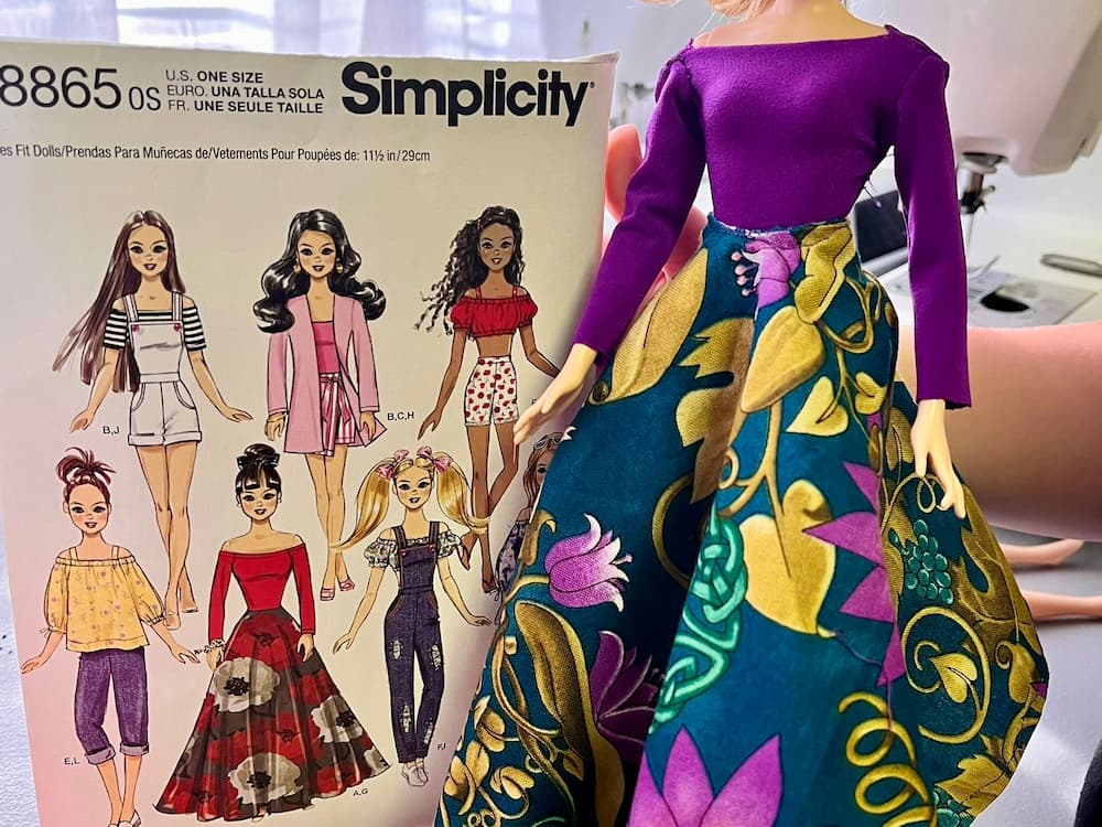 A handmade doll outfit with a bold patterned skirt and purple top displayed next to a Simplicity sewing pattern for doll clothes.