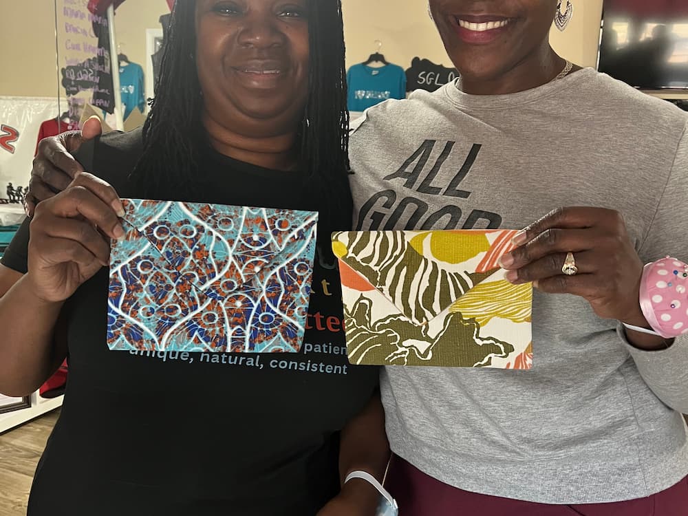 Two smiling women proudly holding handmade fabric envelope clutches with bold, colorful patterns.