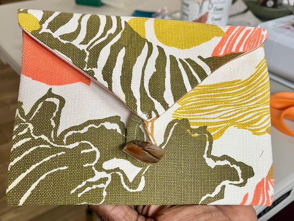 Close-up of a handmade fabric clutch with a button closure, featuring a colorful abstract pattern in green, yellow, orange, and white.
