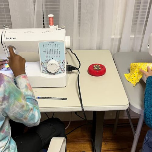 Two young students using sewing machines side by side, working on colorful fabric pieces in a classroom setting.