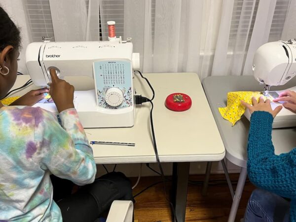 Two young students using sewing machines side by side, working on colorful fabric pieces in a classroom setting.