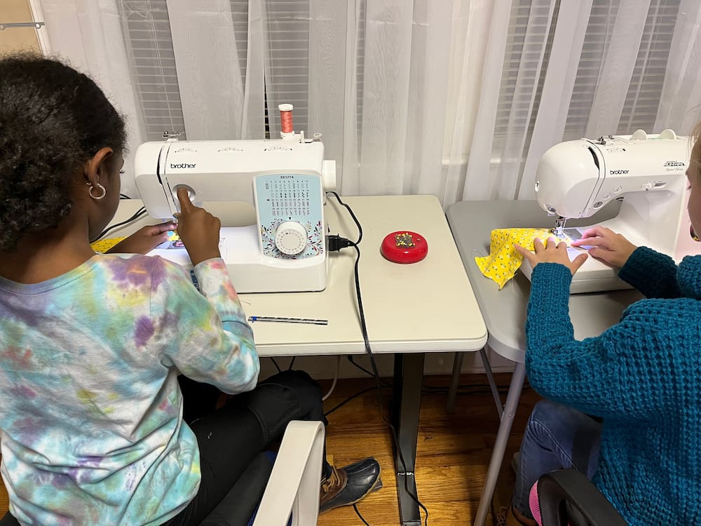 Two young girls using sewing machines to work on fabric projects during a community sewing workshop.