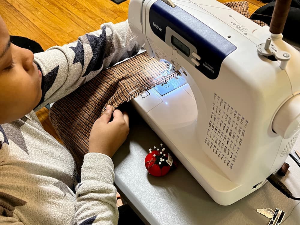 Young student sewing a brown checkered fabric on a Brother sewing machine with a red pincushion nearby.