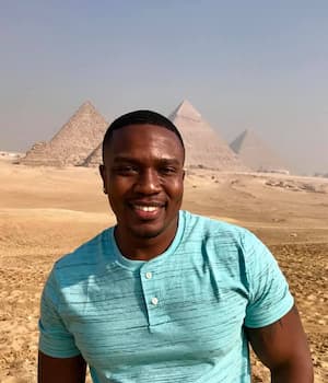 Rously Michel smiling in front of the pyramids in Egypt, wearing a turquoise shirt
