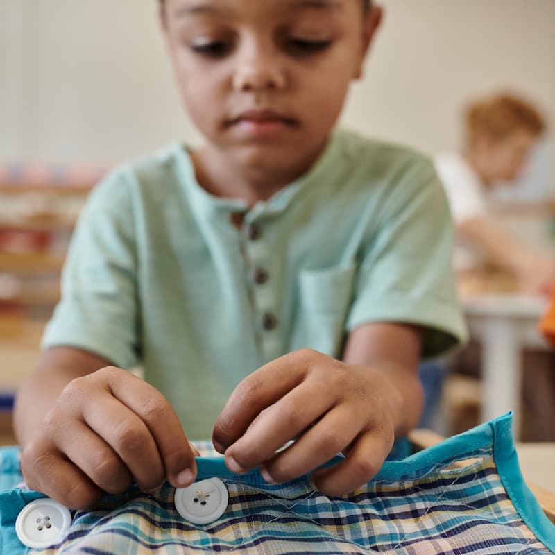 Young boy concentrating on sewing white buttons onto a blue plaid fabric with turquoise trim.