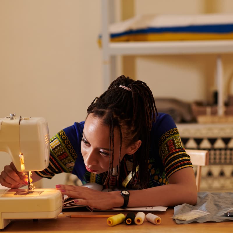 Young woman carefully threading a needle on a sewing machine, with spools of thread on the table.