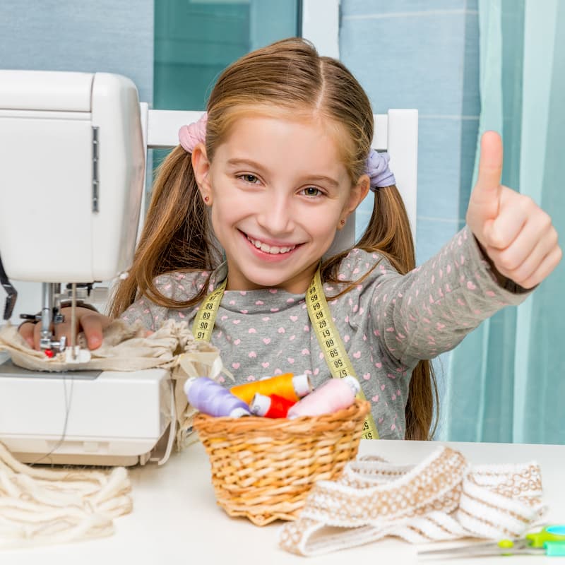 Smiling girl giving a thumbs-up while sitting at a sewing machine with thread spools and fabric on the table.