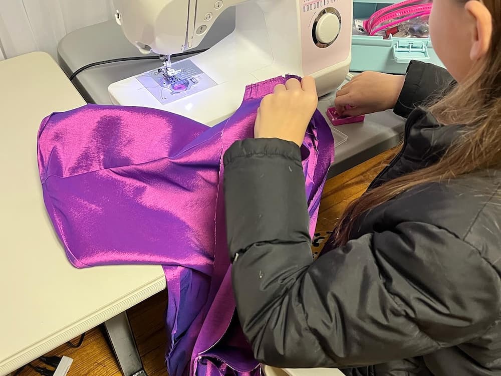 Child sewing a shiny purple fabric using a Brother sewing machine in a classroom setting.