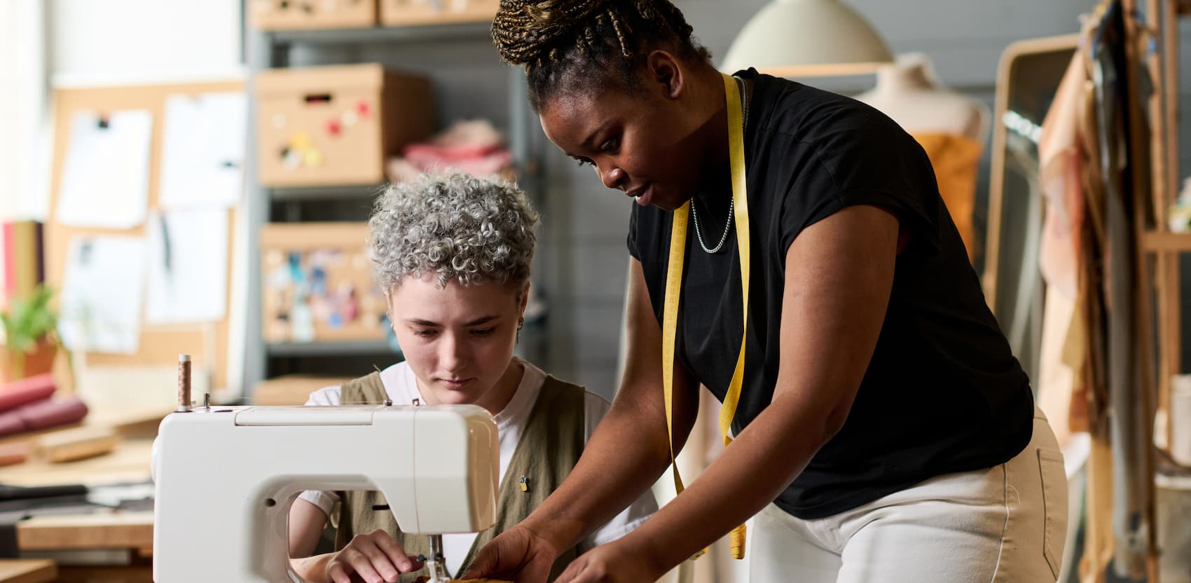 A Black woman volunteer teaching a student to use a sewing machine in a creative studio space.
